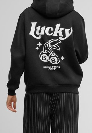 Personne portant un sweat à capuche noir avec le texte « Lucky », deux cerises avec le chiffre 8, et « Good Vibes Only » au dos, assorti à un pantalon à fines rayures.