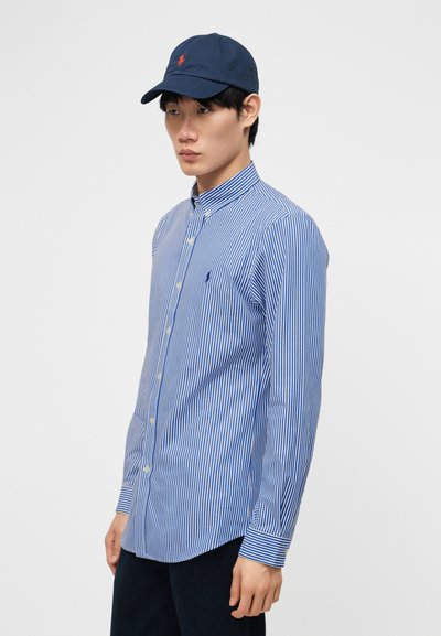 Bărbat tânăr purtând o cămașă cu nasturi în dungi albastre și albe și o șapcă de baseball albastru închis, stând pe un fundal simplu, deschis la culoare.