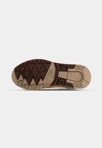 Trail shoe outsole, featuring a brown rubber traction pattern, beige midsole, and textured geometric designs for improved grip.