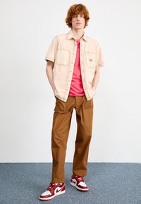 Beige overhemd met korte mouwen en knopen, met twee borstzakken, gecombineerd met een roze T-shirt en bruine recht vallende broek. Rode en witte sneakers.