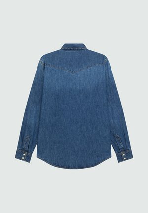 Back view of a long-sleeve blue denim shirt with pointed yoke stitching and buttoned cuffs on a light gray background.