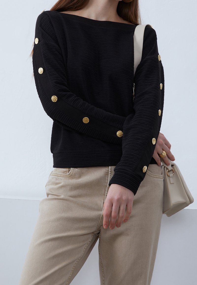 Femme portant un haut noir texturé à manches longues avec des boutons dorés sur les manches, un pantalon beige, et portant un sac à main beige à l'épaule.