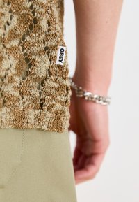 Løsstrikket, beige heklet topp med et teksturert mønster og en hvit "OBEY"-lapp på siden. Bærers over lys grønn, tettsittende stoff.