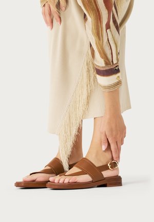 Persona con sandalias planas de cuero marrón con hebilla en el tobillo, falda crema con flecos y camisa de manga larga a rayas, ajustándose la correa de la sandalia.