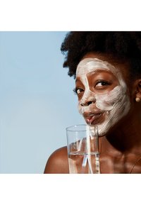 Kvinne med mørk hud og krøllete hår, iført en hvit ansiktsmaske, drikker fra et klart glass med sugerør. Bakgrunnen er lys blå.