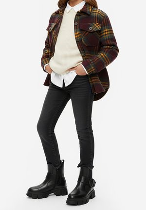 Osoba ubrana w brązową, żółtą i zieloną kratowaną kurtkę na kremowym swetrze i białej koszuli, czarne dżinsy i czarne buty na klamrę.