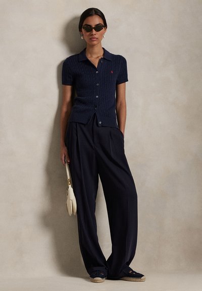 Kobieta w granatowym, krótkim sweterku na guziki, luźnych czarnych spodniach, ciemnych okularach przeciwsłonecznych, espadrylach, trzymająca małą białą torebkę, stoi na tle gładkiej ściany.