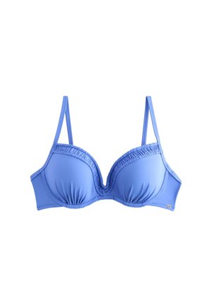 Frau mit langen dunklen Haaren, die einen blauen Bikini trägt und an einem Sandstrand mit Felsen und Meer im Hintergrund steht.