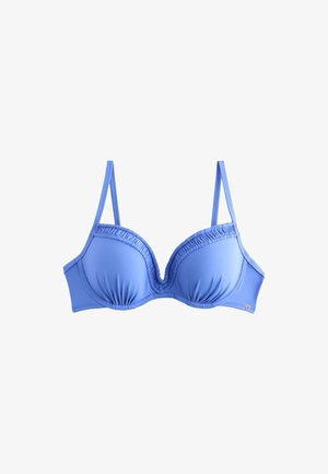 Frau mit langen dunklen Haaren, die einen blauen Bikini trägt und an einem Sandstrand mit Felsen und Meer im Hintergrund steht.