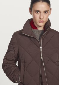 Braune gesteppte Jacke mit hohem Kragen und silbernem Reißverschluss. Das Innenfutter hat eine rote Fütterung, und die Ärmelenden sind mit einem strukturierten Akzent versehen.