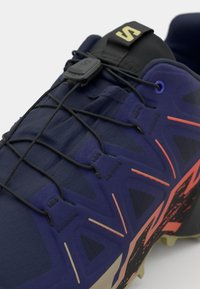 Navy athletic shoe with a textured surface, featuring black elastic laces, orange detailing, a patterned outsole, and a reinforced toe.