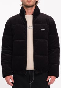 Schwarze Cord-Puffermantel mit einem gesteppten Muster, Reißverschluss, hohem Kragen und Logo auf der Brust, kombiniert mit einem beigen Oberteil darunter.