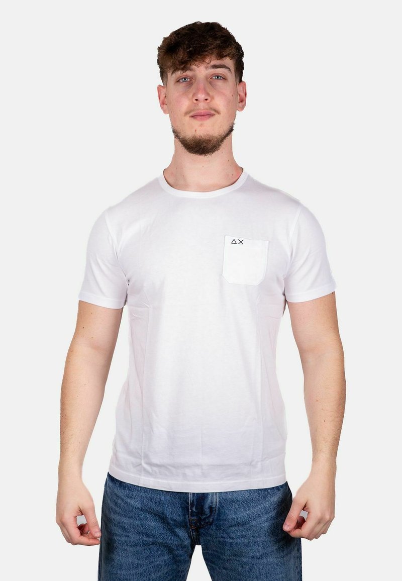 T-shirt bianco a maniche corte in cotone, con una tasca sul petto sinistro con un logo nero e collo a giro. Indossato con jeans blu.