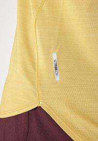 Top deportivo amarillo con un tejido texturizado, parte inferior en color burdeos contrastante y un logo "ACTIBREEZE" en una etiqueta blanca.