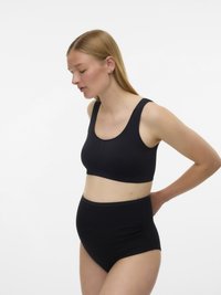 Noseča ženska, oblečena v črne maternitetne spodnje perilo in modrček, stoji v profilu, z eno roko, ki se naslanja na spodnji del hrbta.