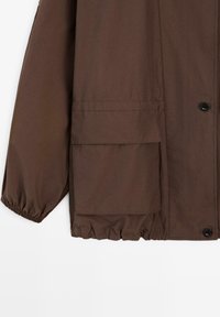 Brązowa kurtka z dużą kieszenią z patką, zapięciem na guziki, elastycznymi mankietami i marszczonym dołem.