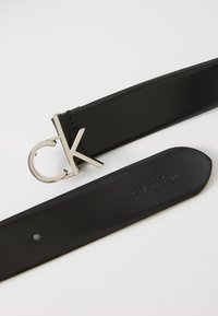 Must mustriba mustra, mille hõbedase tooni metallist pandla ja "CK" logoga. Sujuv tekstuur, millel on subtilne bränding vööl.