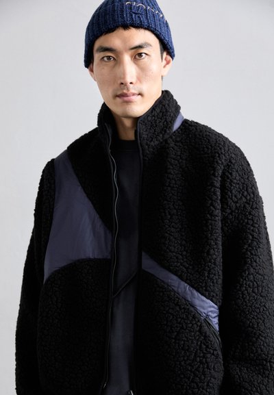 Giacca in pile nera con finitura testurizzata, caratterizzata da un design a pannello contrastante blu navy. Chiusura con zip, colletto alto e tasche laterali.