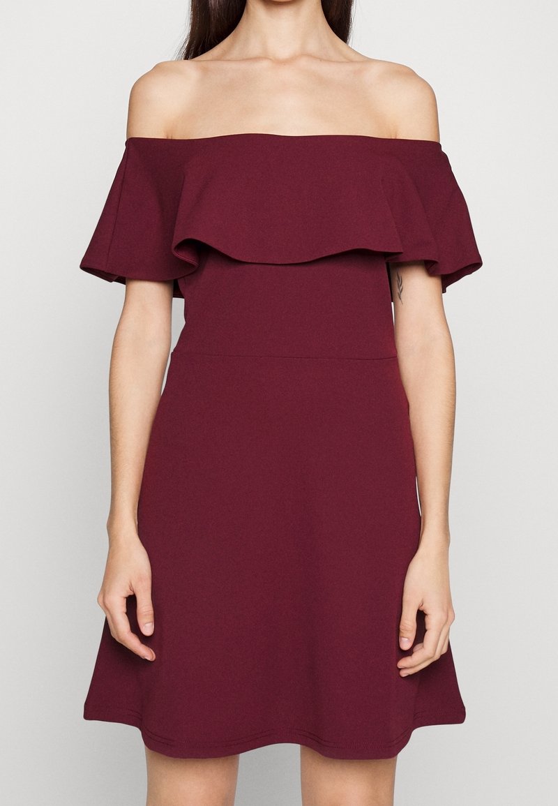 Femme portant une robe bordeaux sans manches et à épaules dénudées, avec un col à volants, debout devant un fond clair uni.