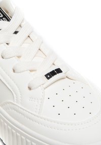 Białe sneakersy z skórzaną cholewką, perforowanym przodem oraz płaskimi sznurowadłami. Posiadają czarną metkę z logo na języku oraz teksturowaną gumową podeszwę.