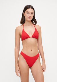 Crveni bikini od strukturirane tkanine; gornji dio ima trokutasti oblik s vezicama oko vrata, a donji dio ima visoki izrez s debelim pojasom.
