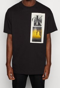 Schwarzes Baumwoll-T-Shirt mit einem rechteckigen Grafikdruck in Weiß und Gelb, auf dem "CK" und "Calvin Klein Jeans" steht. Kurze Ärmel, Rundhalsausschnitt.
