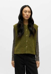 Zelená pletená vesta s kulatým výstřihem, bez rukáv�ů a zlatými knoflíky. Nositá přes šedou košili s dlouhým rukávem a spárována s černými kalhotami.