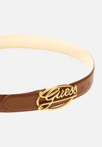 Rudas odinis diržas su auksinės spalvos "Guess" logo sagtimi. Savybės: lygus paviršius, vienas segtukas ir reguliuojamos skylutės.