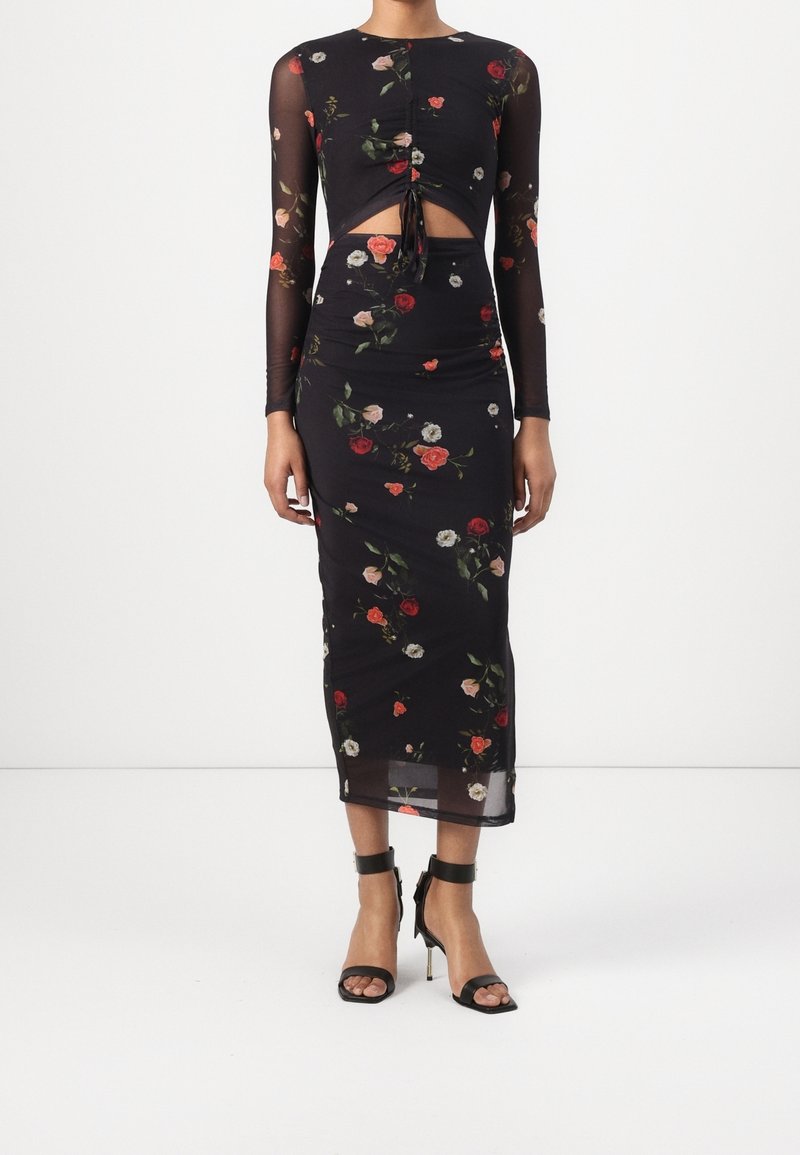 Femme portant une robe midi noire à fleurs avec des manches longues transparentes, une découpe à l'avant nouée à la taille, associée à des talons hauts noirs avec bride à la cheville.