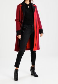 Röd och svart kappa med lång skärning, blixtlås framtill och kontrasterande krage. Bärs över en svart topp och svarta jeans med passform och nitar. Svarta ankelskor.