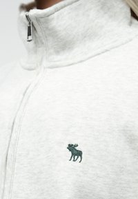 Pull-over en polaire gris clair avec un col haut, affichant un logo d'élan brodé vert foncé à l'avant, et texture douce.