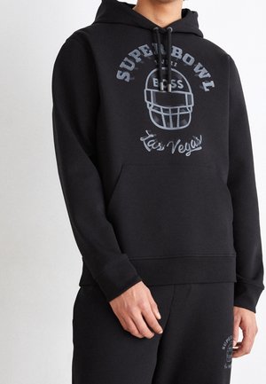 Personne portant un sweat à capuche noir et un pantalon assorti avec l'inscription « Superbowl Las Vegas » et un graphisme de casque de football en gris sur le devant.