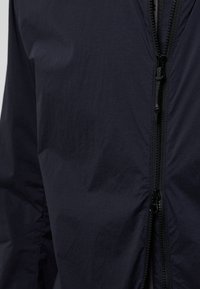 Marineblaue Zip-Jacke mit einer glatten, matten Textur, ausgestattet mit einem zentralen Reißverschluss und klaren Linien im Stoff ohne zusätzliche Muster oder Akzente.