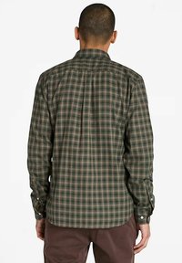 Uomo che indossa una camicia a maniche lunghe a scacchi verde e marrone e pantaloni marroni, in piedi con la schiena rivolta verso l'osservatore su uno sfondo bianco.