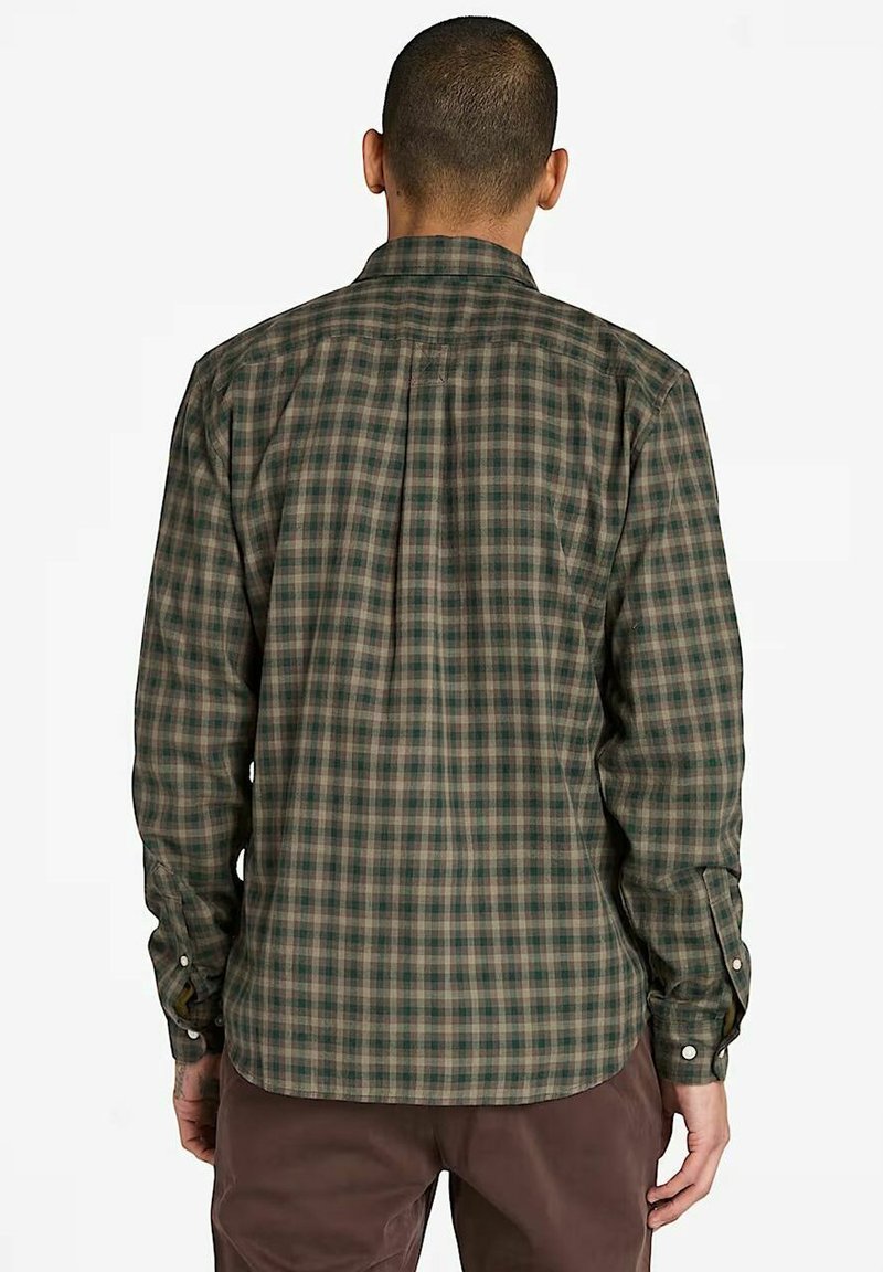 Homme portant une chemise à carreaux verte et marron à manches longues et un pantalon marron, se tenant de dos face au spectateur sur un fond blanc.