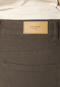 Denim cinza escuro com um remendo de couro castanho exibindo "VERO MODA DENIM." Apresenta costura padrão e um bolso traseiro.