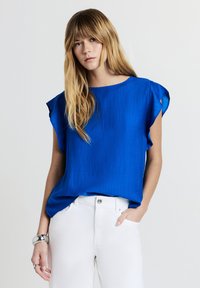 Giovane donna con lunga chioma bionda indossa un blusa azzurra a maniche corte, infilata in pantaloni bianchi, accessoriate con bracciale e anello d'argento.