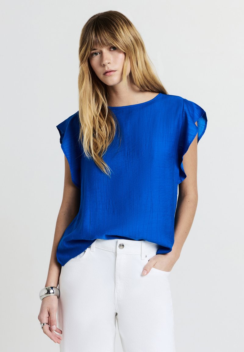 Giovane donna con lunga chioma bionda indossa un blusa azzurra a maniche corte, infilata in pantaloni bianchi, accessoriate con bracciale e anello d'argento.