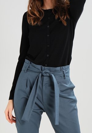 Frau trägt ein schwarzes, knopfbewehrtes Langarmshirt und hoch taillierte blaue Hosen, vorne mit einem breiten Stoffgürtel gebunden.