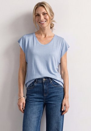 Uśmiechnięta kobieta o blond włosach, ubrana w jasnoniebieską koszulę z krótkim rękawem i niebieskie dżinsy, stojąca na tle gładkiej, jasnej ściany.