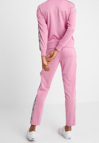 Rosa Trainingsanzug-Set mit Reißverschlussjacke und schmal zulaufender Hose. Schwarze Seitenstreifen mit Akzenten an Bündchen und Knöcheln. Glatter Stoff.