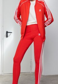 Červená sportovní bunda s bílými pruhy a logem, spárovaná s odpovídajícími červenými legínami s bílými postranními pruhy. Hladká textura materiálu.