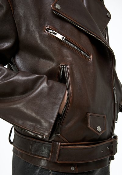 Sötétbarna bőr dzseki ezüst cipzárokkal, deréknál övvel és egy kéz félig a oldalsó cipzáras zsebben.