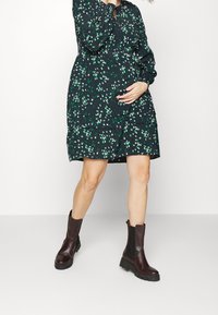 Vestido de estampado floral en azul marino con flores verdes y blancas, con un diseño en capas. Combinado con botas de tobillo burdeos y una silueta esbelta.