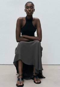 Top negro sin mangas combinado con una falda gris fluida. La modelo lleva sandalias negras con tiras en el tobillo, sentada contra un fondo simple.