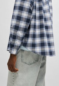 Rutigt bomullsskjorta i marinblått, grått och vitt. Med långa ärmar, knappmanschett och rundad kant. Kombinerad med ljusgrå jeans.