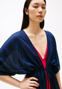 Cămașă din voal de marină, stil kimono, cu mâneci voluminioase, talie strânsă și decolteu adânc în V, purtată peste un costum de baie roz.
