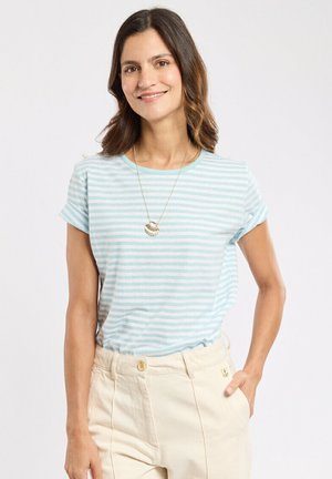 Femme souriante, portant un t-shirt à rayures bleu clair et blanc, un pantalon beige taille haute et un collier pendentif coquillage doré, debout devant un fond blanc.