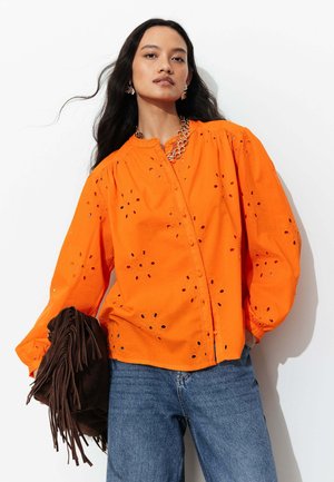 Femme aux longs cheveux foncés porte un chemisier orange à œillets, un jean bleu, un collier chaîne en or et tient un sac à franges marron sur un fond blanc.