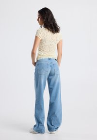 Woman with dark hair wearing a pale yellow lace short-sleeve top, light blue wide-leg jeans, and white sneakers, standing against a plain background.